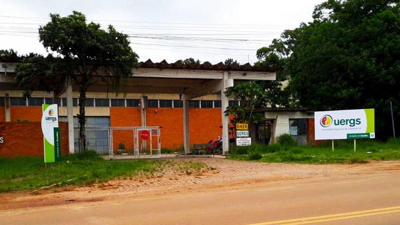 Ao fundo, pavilhão de tijolos à vista e um portão grande de entrada. À frente, o totem de identificação da Uergs à esquerda e placa da Uergs à direita.