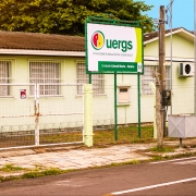 Foto do prédio da Unidade, de apenas 1 andar e de paredes em tom de verde claro. À frente, junto à cerca, a placa de identificação da Uergs.