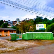 Em primeiro plano, um muro de tijolos à vista pintados de verde, com uma placa de identificação da Uergs, à direita. Ao fundo, o prédio da Unidade, pintado de laranja.