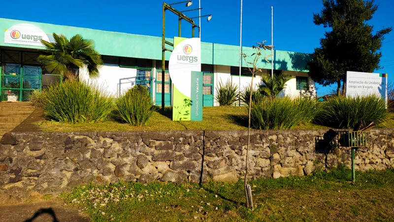 À frente, muro baixo de pedras contém uma área de jardim. Ao fundo, totem da unidade e o prédio térreo.