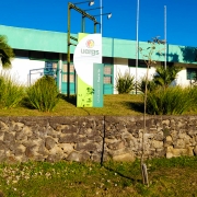 À frente, muro baixo de pedras contém uma área de jardim. Ao fundo, totem da unidade e o prédio térreo.