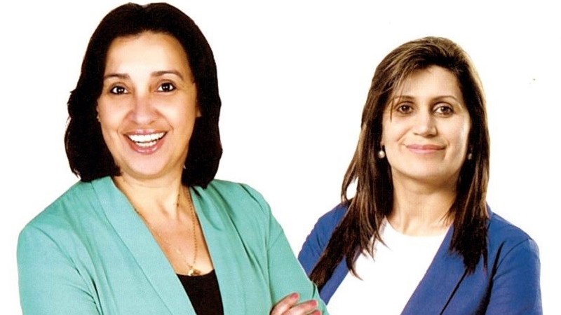 Duas mulheres de pele clara e sorridentes. A da esquerda tem cabelos castanho escuros, lisos, na altura dos ombros e veste blusa preta e terno verde claro. A da direita tem cabelos louros, lisos e compridos, e veste blusa branca e terno azul.