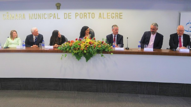 Palco com sete pessoas sentadas à mesa, que contém um arranjo floral ao centro. Ao fundo, lê-se Câmara Municipal de Porto Alegre.