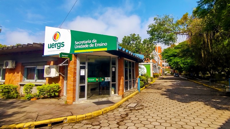 Foto colorida de um prédio de 1 andar, com paredes de tijolos à vista, telhado azul e portas de vidro. Na fachada, uma placa verde onde se lê Secretaria da Unidade de Ensino. Ao lado esquerdo, o logotipo da Uergs.