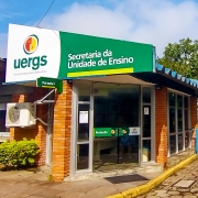 Foto colorida de um prédio de 1 andar, com paredes de tijolos à vista, telhado azul e portas de vidro. Na fachada, uma placa verde onde se lê Secretaria da Unidade de Ensino. Ao lado esquerdo, o logotipo da Uergs.