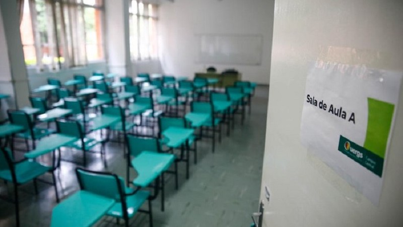 Foto de sala de aula. À direita, uma porta aberta contendo um cartaz de identificação da sala. À esquerda, em segundo plano, cadeiras universitárias dispostas em fileiras.