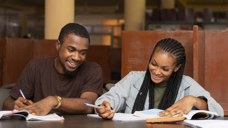 Dois jovens negros sorridentes (um homem e uma mulher), sentados olhando para livros e cadernos, com canetas nas mão. Ele tem cabelos curtos, bigode e barba, e veste camiseta marrom. Ela tem cabelos compridos com tranças nagô e veste jaqueta jeans.