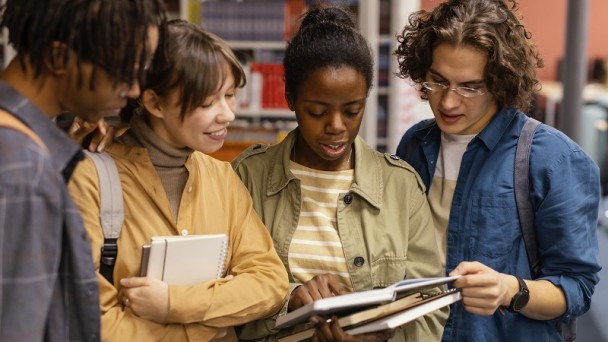 Quatro pessoas jovens conversam e olham um livro em uma biblioteca.