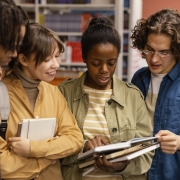 Quatro pessoas jovens conversam e olham um livro em uma biblioteca.
