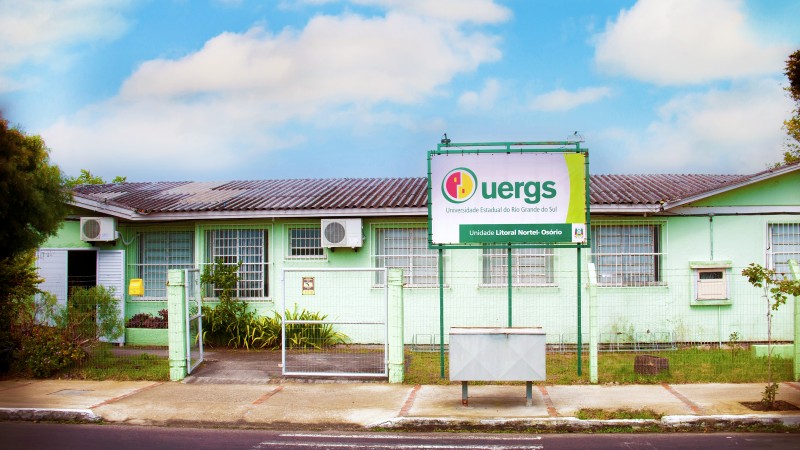 Foto do prédio da Unidade, de apenas 1 andar e de paredes em tom de verde claro. À frente, junto à cerca, a placa de identificação da Uergs.