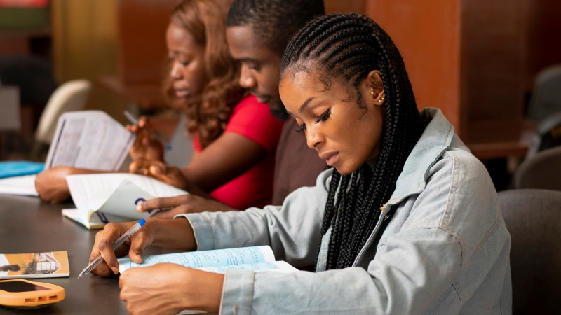 Foto de três estudantes negros sentados lado a lado, olhando atentamente para livros, com canetas nas mãos. A foto é da visão lateral deles, sendo duas mulheres nas pontas e um homem ao centro.