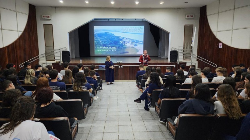 Foto colorida de um auditório. As fileiras de cadeiras estão ocupadas por estudantes posicionados de frente para um palco, do qual um homem e uma mulher estão próximos e em pé. Ao fundo, há um telão que exibe uma cena do documentário.