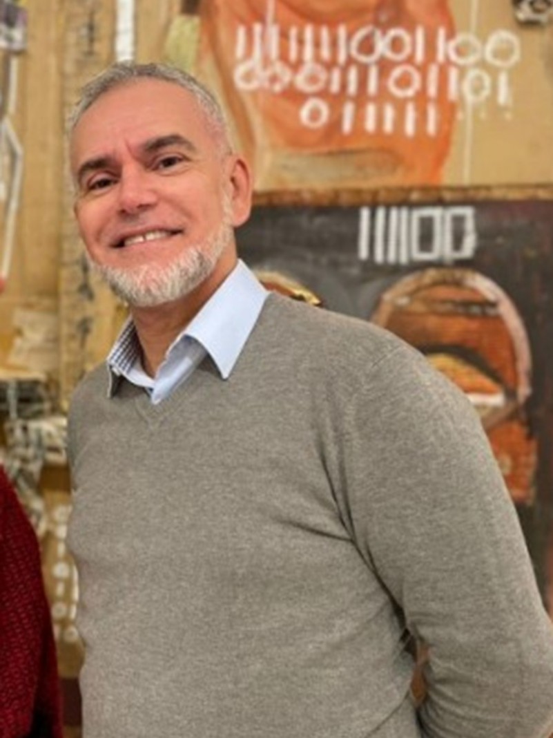 Homem de pele clara, cabelos e barba grisalhos, ilhos escuros. Sorri para a foto. Veste camista clara por baixo de um sueter cinza.