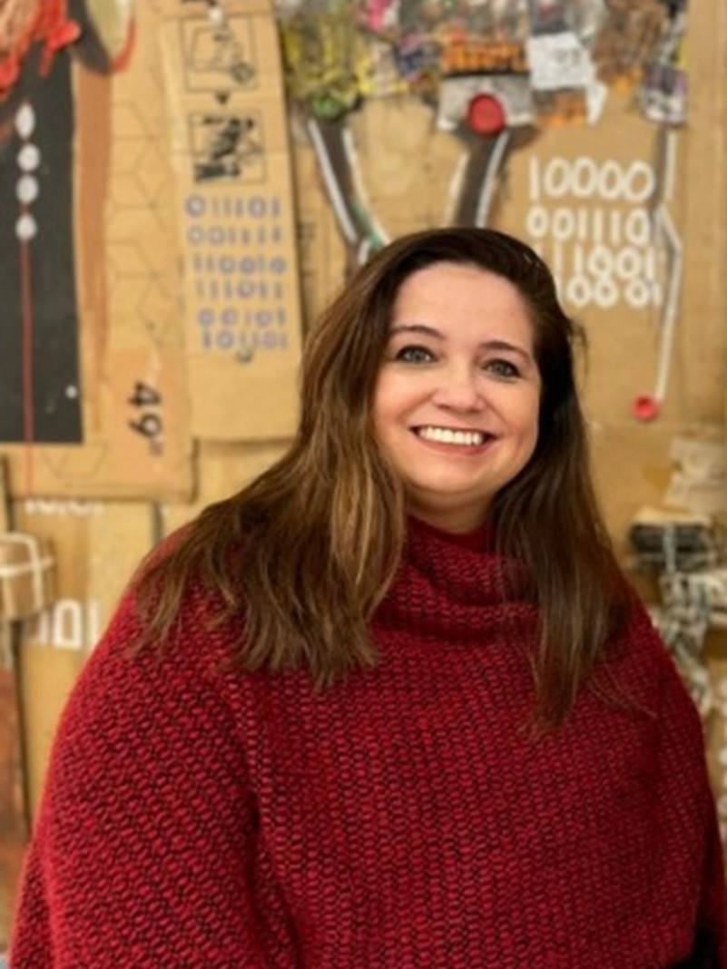 Mulher de pele clara, cabelos longos castanhos, olhos escuros. Sorri para a foto. Veste poncho vermelho em trico.