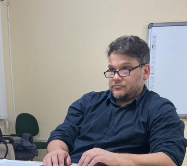 Homem de cabelos e barba grisalhos, usando óculos de grau, com semblante sério, sentado a uma mesa com um quadro branco ao fundo.