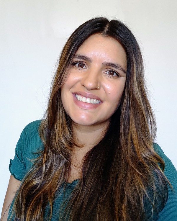 Foto colorida de uma mulher sorridente, morena clara, de olhos castanhos, cabelos longos castanhos com mechas, vestindo uma roupa verde.