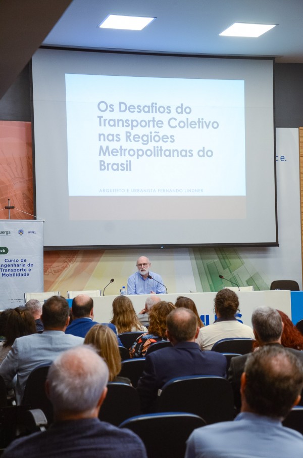 Foto colorida. Foto que mostra pessoas de costas, sentadas em poltronas e, à frente delas, o palestrante do evento sentado a uma mesa. Atrás e acima do palestrante, um telão com o slide da palestra.
