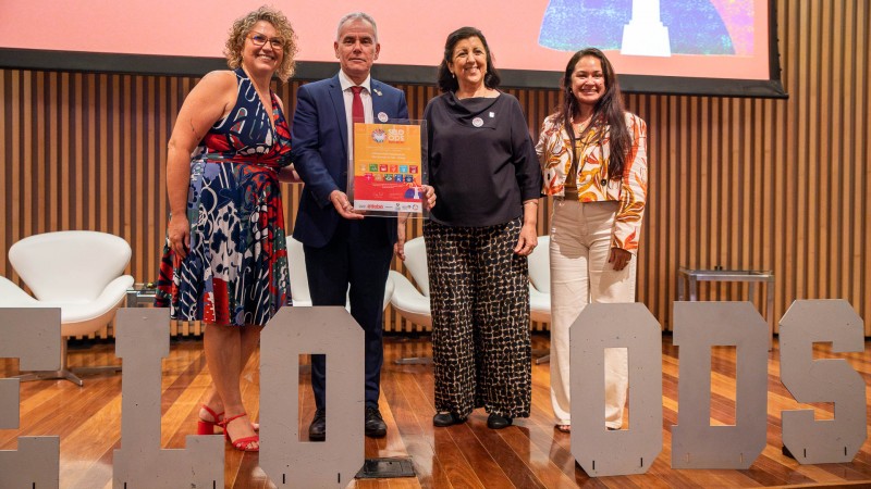 Foto de quatro pessoas sorridentes, sendo três mulheres e um homem. Eles estão em um palco com placas de letras brancas à sua frente, escrito "SELO ODS". O homem segura uma placa com a certificação do Selo ODS.