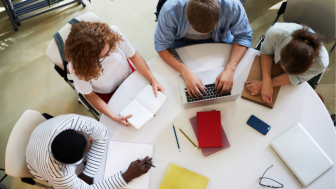 Foto colorida, vista de cima, de quatro estudantes estudando ao redor de uma mesa branca, com cadernos e um notebook.