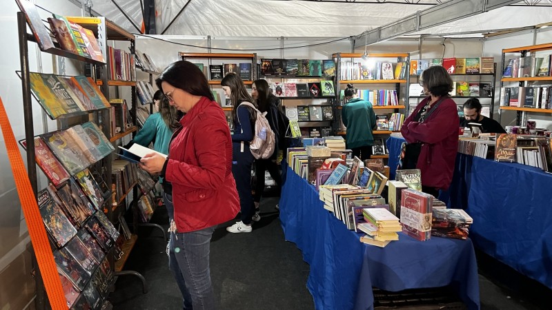 Foto colorida de ambiente com diversas prateleiras verticais com livros e mesas ao centro, cobertas com tecido azul e também com muitos livros. Há pessoas com livros nas mãos e também olhando as prateleiras.