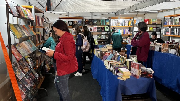 Foto colorida de ambiente com diversas prateleiras verticais com livros e mesas ao centro, cobertas com tecido azul e também com muitos livros. Há pessoas com livros nas mãos e também olhando as prateleiras.