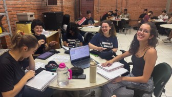 Foto de uma sala de aula onde estudantes estão organizados(as) em grupos em mesas redondas. Em primeiro plano um grupo de jovens que sorriem e interagem.