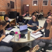 Foto de uma sala de aula onde estudantes estão organizados(as) em grupos em mesas redondas. Em primeiro plano um grupo de jovens que sorriem e interagem.