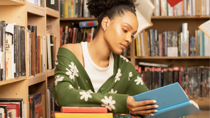 Foto de uma mulher jovem negra, de cabelos presos, vestindo regata branca com um casaco verde com flores brancas por cima. Ela está sentada segurando um livro de capa azul, enquanto olha para ele. Ao fundo, prateleira com diversos livros.