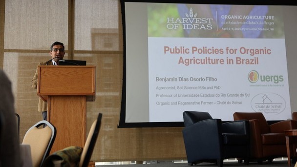 Foto de uma pessoa em pé sobre um palco, palestrando apoiado em um púlpito. À sua esquerda, há um painel com o título da palestra em Inglês “Public Policies for Organic Agriculture in Brazil”, informações sobre o palestrante e o logotipo da Uergs. . 