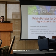 Foto de uma pessoa em pé sobre um palco, palestrando apoiado em um púlpito. À sua esquerda, há um painel com o título da palestra em Inglês “Public Policies for Organic Agriculture in Brazil”, informações sobre o palestrante e o logotipo da Uergs. . 