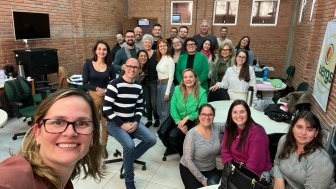 Foto colorida tirada em ambiente interno, numa sala com paredes de tijolos à vista e três janelas pequenas na parte superior. O espaço tem piso branco e é mobiliado com cadeiras giratórias e mesas redondas brancas.
No centro da imagem, há um grupo de aproximadamente 25 pessoas, adultas, a maioria aparentando estar entre os 30 e 60 anos, posando sorridentes para a foto. Estão vestidas de forma casual, com roupas em tons variados como verde, cinza, rosa, preto, branco e azul. 
No canto esquerdo inferior da imagem, uma mulher de óculos e cabelos loiros escuros na altura dos ombros tira a selfie. Ela está sorrindo e veste uma blusa marrom.
Ao fundo, à direita, há uma faixa com o logotipo da Uergs.
