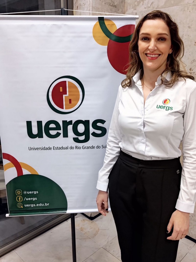 Mulher sorridente de pele cara, olhos castanhos e cabelo louro de tamanho médio e levemente ondulado. Veste camisa branca, com logotipo da Uergs bordado, localizado no lado esquerdo do peito, e calça preta. Atrás dela, um banner com o logotipo da Uergs sobre fundo branco.