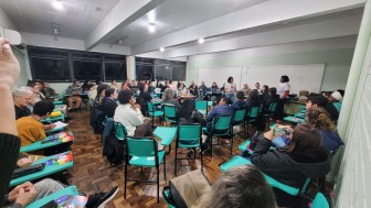 A foto mostra uma sala de aula ampla e bem iluminada, com piso de madeira e paredes de tijolos pintados de verde claro. Cerca de 60 pessoas participam de uma roda de conversa, dispostas em cadeiras escolares com mesas acopladas, organizadas em formato circular e semicircular. A maioria das cadeiras é verde água.
No centro da sala, duas mulheres de camiseta branca falam para o grupo. Uma está em pé, próximo ao quadro branco, e outra ao centro da roda, segurando um leque com a mesma identidade visual da camiseta.
O público é diverso, com pessoas de diferentes faixas etárias, tons de pele, estilos e gêneros. Algumas estão atentas às falas, outras leem folhetos ou observam ao redor. Quase todas carregam o leque colorido da campanha sobre igualdade.
Ao fundo, grandes janelas com vidros escuros ocupam a maior parte da parede lateral. 

