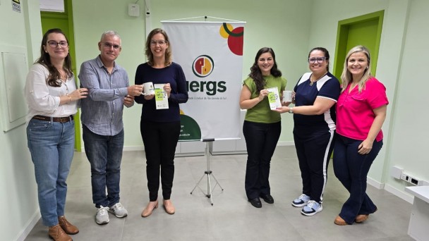 Seis pessoas estão em pé, lado a lado, sorrindo para a câmera em um ambiente interno com paredes verde-claro e portas verdes. No centro do grupo, ao fundo, há um banner com o logotipo da Uergs (Universidade Estadual do Rio Grande do Sul).
Da esquerda para a direita:
A primeira pessoa é uma mulher de cabelos castanhos, soltos e ondulados, usando óculos, blusa branca de mangas bufantes e jeans. Ela está com uma das mãos no ombro do homem ao lado.


O segundo é um homem de cabelo grisalho curto, usando óculos, camisa azul-clara listrada e calça jeans. Ele segura uma caneca branca junto com a terceira pessoa.


A terceira é uma mulher loira, de óculos, usando blusa azul-escura e calça preta. Ela segura a caneca e um panfleto.


A quarta é uma mulher de cabelos castanhos escuros e longos, usando uma blusa verde e calça preta. Ela segura o mesmo tipo de panfleto.


A quinta é uma mulher de óculos com armação azul, cabelos presos, vestindo uma camiseta azul com branco e calça esportiva. Ela segura um copo transparente.


A sexta é uma mulher de cabelos loiros, soltos, vestindo uma blusa rosa-choque e calça jeans escura. Ela está com a mão nas costas da colega ao lado.


O ambiente é iluminado por luz artificial, com piso cinza claro e teto de placas modulares. Todos parecem felizes e participando de um momento comemorativo ou de entrega simbólica de brindes.
