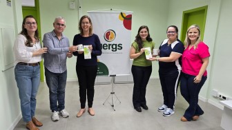 Seis pessoas estão em pé, lado a lado, sorrindo para a câmera em um ambiente interno com paredes verde-claro e portas verdes. No centro do grupo, ao fundo, há um banner com o logotipo da Uergs (Universidade Estadual do Rio Grande do Sul).
Da esquerda para a direita:
A primeira pessoa é uma mulher de cabelos castanhos, soltos e ondulados, usando óculos, blusa branca de mangas bufantes e jeans. Ela está com uma das mãos no ombro do homem ao lado.


O segundo é um homem de cabelo grisalho curto, usando óculos, camisa azul-clara listrada e calça jeans. Ele segura uma caneca branca junto com a terceira pessoa.


A terceira é uma mulher loira, de óculos, usando blusa azul-escura e calça preta. Ela segura a caneca e um panfleto.


A quarta é uma mulher de cabelos castanhos escuros e longos, usando uma blusa verde e calça preta. Ela segura o mesmo tipo de panfleto.


A quinta é uma mulher de óculos com armação azul, cabelos presos, vestindo uma camiseta azul com branco e calça esportiva. Ela segura um copo transparente.


A sexta é uma mulher de cabelos loiros, soltos, vestindo uma blusa rosa-choque e calça jeans escura. Ela está com a mão nas costas da colega ao lado.


O ambiente é iluminado por luz artificial, com piso cinza claro e teto de placas modulares. Todos parecem felizes e participando de um momento comemorativo ou de entrega simbólica de brindes.
