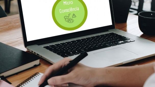 Sobre uma mesa, um notebook, na tela o logotipo do projeto MicroConsciência. A mão de uma pessoa segura uma caneta repousando sobre um bloco de anotações.