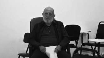 Foto em preto e banco de um homem idoso, calvo, com barba branca. Ele está sério,  sentado em uma sala de aula e com folhas de papel nas mãos.