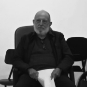 Foto em preto e banco de um homem idoso, calvo, com barba branca. Ele está sério,  sentado em uma sala de aula e com folhas de papel nas mãos.