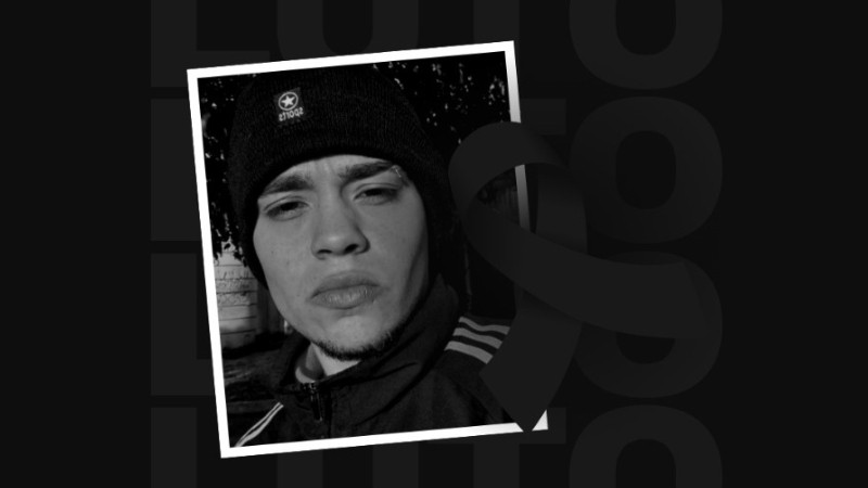 Foto em preto e branco de um jovem sério, moreno claro, de sobrancelhas e lábios espessos, vestindo jaqueta e uma touca de lã pretas. Sobre a foto, há a imagem de um laço preto, simbolizando luto.