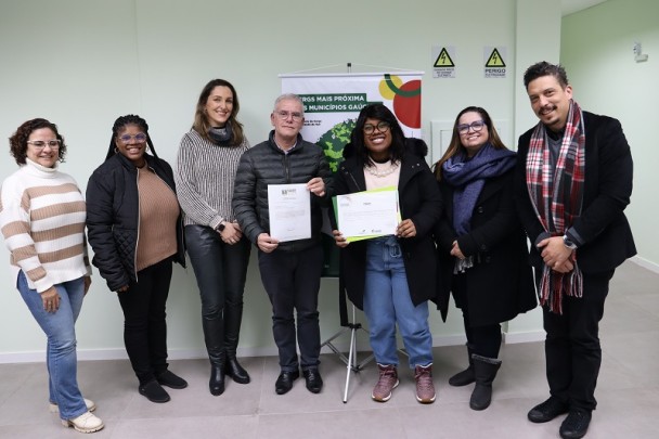 Em um ambiente interno com paredes claras, oito pessoas posam sorridentes para a foto em pé, lado a lado. Ao centro, duas pessoas seguram documentos: um homem de cabelo grisalho, vestindo óculos, jaqueta preta e calça escura; ao seu lado, uma mulher negra com cabelos longos e soltos, usando óculos, colar de pérolas, blusa clara, jaqueta preta, calça jeans e tênis rosa. Atrás deles, há um banner com o mapa da Uergs e os dizeres “Uergs mais próxima dos municípios gaúchos”. À esquerda da imagem, três mulheres estão lado a lado, usando roupas de inverno: a primeira veste suéter listrado em branco e bege, calça jeans e calçado claro; a segunda é negra, com óculos, cabelo com tranças, blusa marrom, jaqueta preta e calça escura; a terceira tem cabelo castanho claro, veste blusa de lã listrada em cinza e bege, cachecol cinza e calça preta. À direita da mulher que segura um documento, estão mais duas pessoas: uma mulher com cabelo castanho, óculos, cachecol azul e casaco preto, e um homem com cabelo escuro, bigode e barba, vestindo casaco preto, calça escura e cachecol xadrez em vermelho, cinza e branco. Ao fundo, há duas placas amarelas de “perigo – eletricidade” fixadas na parede.
