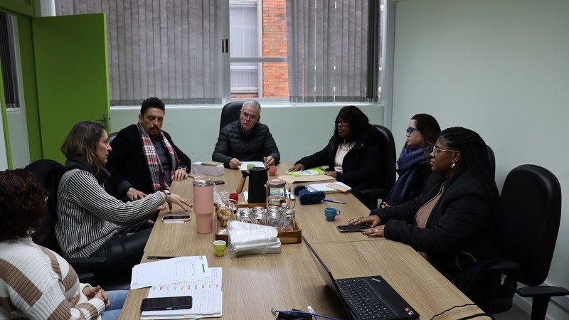 Em uma sala de reuniões com paredes claras e janelas com persianas cinzas parcialmente fechadas, sete pessoas estão sentadas ao redor de uma mesa retangular de madeira clara. A maioria delas está usando roupas de inverno. À esquerda, três mulheres e um homem estão sentados de um lado da mesa, enquanto, do outro lado, há três pessoas – duas mulheres e um homem. A mulher que está falando gesticula com as mãos, enquanto os demais a observam atentamente, em clima de escuta e concentração.
Sobre a mesa, há diversos objetos: cadernos, papéis, celulares, canecas coloridas, potes de vidro, uma garrafa térmica e bandeja com biscoitos. Há também um notebook aberto com cabos conectados. A luz natural entra pela janela ao fundo, e a porta verde da sala está entreaberta à esquerda da imagem.
