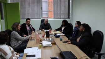 Em uma sala de reuniões com paredes claras e janelas com persianas cinzas parcialmente fechadas, sete pessoas estão sentadas ao redor de uma mesa retangular de madeira clara. A maioria delas está usando roupas de inverno. À esquerda, três mulheres e um homem estão sentados de um lado da mesa, enquanto, do outro lado, há três pessoas – duas mulheres e um homem. A mulher que está falando gesticula com as mãos, enquanto os demais a observam atentamente, em clima de escuta e concentração.
Sobre a mesa, há diversos objetos: cadernos, papéis, celulares, canecas coloridas, potes de vidro, uma garrafa térmica e bandeja com biscoitos. Há também um notebook aberto com cabos conectados. A luz natural entra pela janela ao fundo, e a porta verde da sala está entreaberta à esquerda da imagem.
