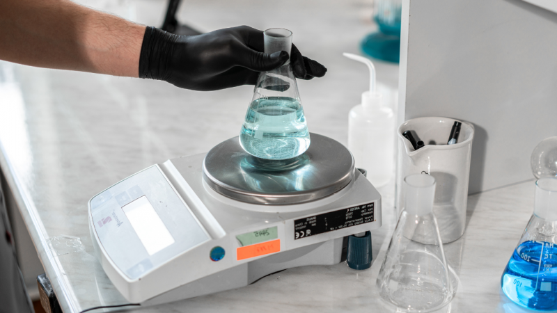 Sobre uma bancada há uma balança de precisão em que uma pessoa apoia uma vidraria com líquido dentro. Ao redor, vidrarias e utensílios de laboratório.