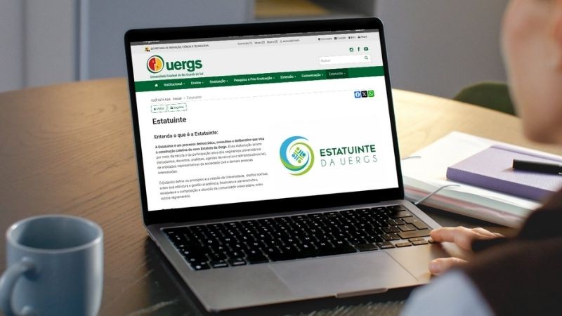 **Audiodescrição da imagem:**

Pessoa vista parcialmente, do lado direito da imagem, está sentada à mesa em ambiente interno e utiliza um notebook. Na tela do computador, está aberta a página da Uergs (Universidade Estadual do Rio Grande do Sul), com o título “Estatuinte” em destaque. Abaixo do título, há um breve texto explicando o processo da Estatuinte, seguido do logotipo colorido da Estatuinte da Uergs, composto por formas circulares e figuras humanas estilizadas.

Sobre a mesa de madeira clara, há uma caneca branca à esquerda do notebook, e ao fundo, um caderno roxo com folhas, uma caneta e um cabo branco enrolado. O ambiente tem iluminação suave e tom acolhedor, com uma cadeira verde parcialmente visível no fundo.
