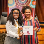Duas mulheres negras estão em pé, sorrindo e posando para a foto em um em um auditório. À esquerda está a deputada Laura Sito, com cabelo cacheado solto, óculos redondos pretos, camisa branca, gravata amarela estampada e saia cinza. Ela segura uma medalha e um certificado com a mulher à sua direita. À direita, a professora Marta Nunes veste um vestido listrado em cores vivas (vermelho, verde, rosa e amarelo) e colar dourado. Ambas seguram o certificado de reconhecimento concedido à professora, com a identidade visual da “Medalha Preta Roza”. Ao fundo, há cadeiras de plenário, madeira revestindo a parede e um banner decorado com mandalas coloridas e os dizeres: “Medalha Preta Roza – A heroína preta do RS – Laura Sito”.
