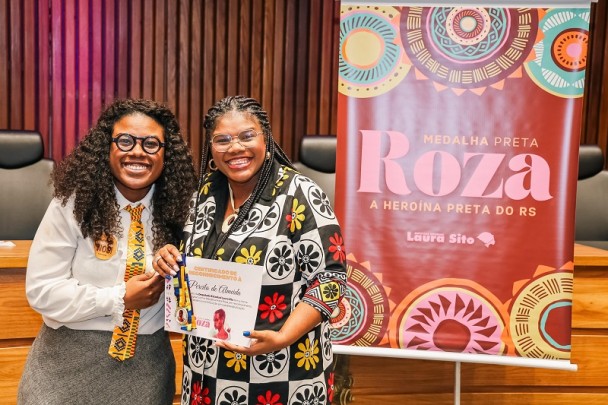 Duas mulheres negras estão em pé, sorrindo, posando para a foto em um auditório. Ambas usam óculos e estão com as mãos entrelaçadas. À esquerda, a deputada Laura Sito veste camisa branca, gravata amarela estampada e saia cinza. À direita, a professora Percila Almeida segura um certificado e uma medalha recebidos em homenagem. Ela veste um vestido preto com estampa colorida de flores grandes em vermelho, amarelo e branco. Ao fundo, um banner com grafismos circulares coloridos traz os dizeres: “Medalha Preta Roza – A heroína preta do RS – Laura Sito”. 
