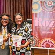 Duas mulheres negras estão em pé, sorrindo, posando para a foto em um auditório. Ambas usam óculos e estão com as mãos entrelaçadas. À esquerda, a deputada Laura Sito veste camisa branca, gravata amarela estampada e saia cinza. À direita, a professora Percila Almeida segura um certificado e uma medalha recebidos em homenagem. Ela veste um vestido preto com estampa colorida de flores grandes em vermelho, amarelo e branco. Ao fundo, um banner com grafismos circulares coloridos traz os dizeres: “Medalha Preta Roza – A heroína preta do RS – Laura Sito”. 
