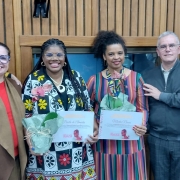 Seis pessoas adultas estão alinhadas lado a lado, posando para a foto em um ambiente interno com paredes de madeira. Ao centro, duas mulheres negras — Percila de Almeida (à esquerda) e Marta Nunes (à direita) — sorriem segurando certificados e vasos com orquídeas, recebidos em homenagem. Percila veste um vestido estampado com flores grandes em preto, branco, vermelho e amarelo, e usa tranças longas. Marta veste um vestido listrado com cores vibrantes. Ao redor delas, quatro pessoas brancas — duas mulheres à esquerda e um homem e uma mulher à direita — as acompanham, também sorrindo. O grupo transmite uma atmosfera de apoio, celebração e reconhecimento.