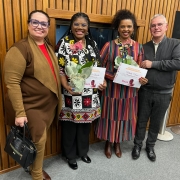 Quatro pessoas adultas estão em pé, lado a lado, sorrindo para a câmera em um ambiente interno de madeira clara. Ao centro estão duas mulheres negras homenageadas: Percila de Almeida (à esquerda), com tranças longas e vestido preto com estampas florais grandes em amarelo, branco e vermelho, e Marta Nunes (à direita), com cabelo preso em coque volumoso e vestido listrado em tons vibrantes. Ambas seguram vasos com orquídeas e certificados com o selo “Medalha Preta Roza”. À esquerda está uma mulher branca com óculos, cabelo preso, vestindo conjunto em tons terrosos e segurando uma bolsa preta. À direita, um homem branco de cabelo grisalho e óculos veste jaqueta cinza e calça xadrez.