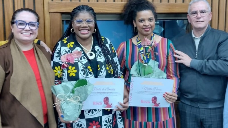 Quatro pessoas estão alinhadas, sorrindo para a câmera em um ambiente interno com paredes de madeira. Ao centro, as professoras negras Percila de Almeida (à esquerda) e Marta Nunes (à direita) seguram certificados de homenagem e vasos com orquídeas embaladas em papel verde. Percila usa um vestido preto com estampa de flores grandes nas cores amarela, branca e vermelha, e tranças longas; Marta veste um vestido listrado verticalmente em tons vivos. À esquerda, uma mulher branca com óculos grandes e roupa em tons terrosos sorri e apoia a mão no ombro de Percila. À direita, um homem branco de cabelo grisalho e óculos apoia a mão nas costas de Marta.